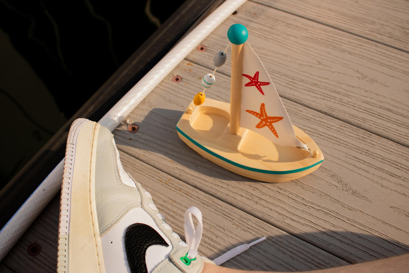 Theme Shot Wooden toy sailboat with starfish sails, colorful bead garland, and partially visible Air Force 1 sneaker on a wooden dock with dark water in the background.