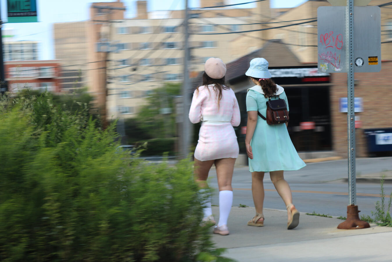 Action Shot Two young women, dressed in fashionable pastel outfits, walk along a city sidewalk. The woman on the left wears a pink plaid skirt suit and a beret, while the woman on the right wears a light blue dress and a bucket hat, and carries a small brown backpack.
