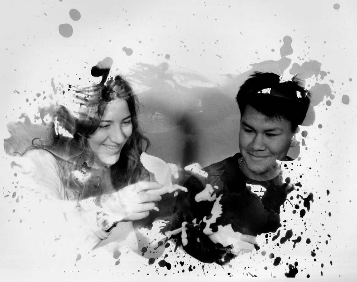 Dark Room Work A black and white photo of two people smiling and playing with paper birds. Ink splatters overlay the scene, adding a creative, joyful feel.