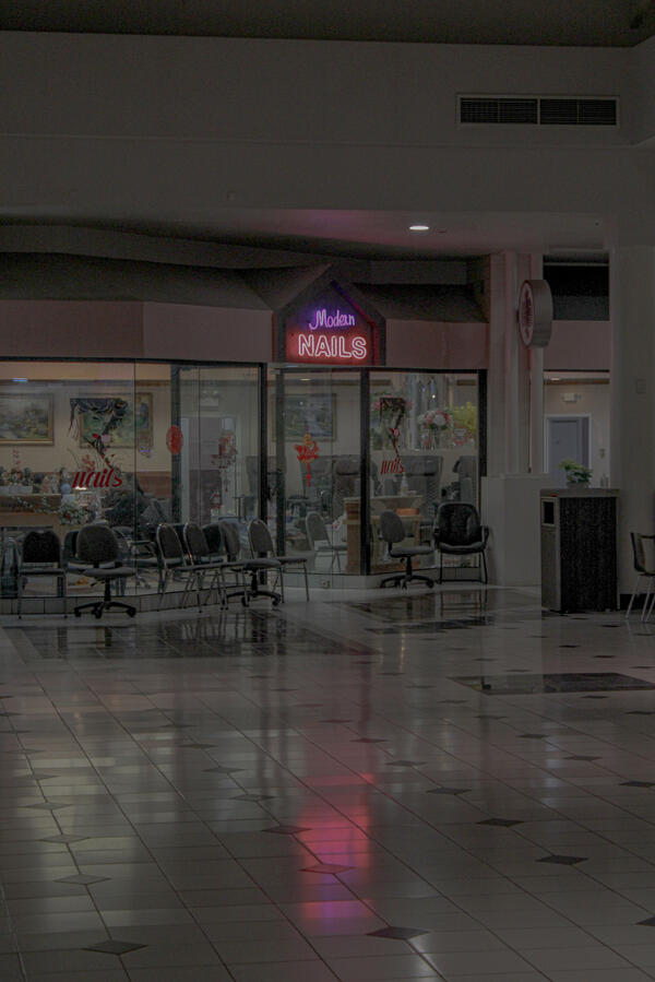 Old Mall Shot Neon sign reading "Modern Nails" above the entrance to a nail salon inside a dimly lit shopping mall, with rows of empty chairs in the waiting area.