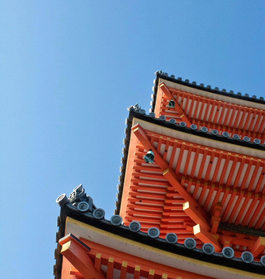 Low-angle view of an ornate orange-and-white Japanese temple roof against a clear blue sky, showing layered eaves, decorative roof tiles, and wooden beams.