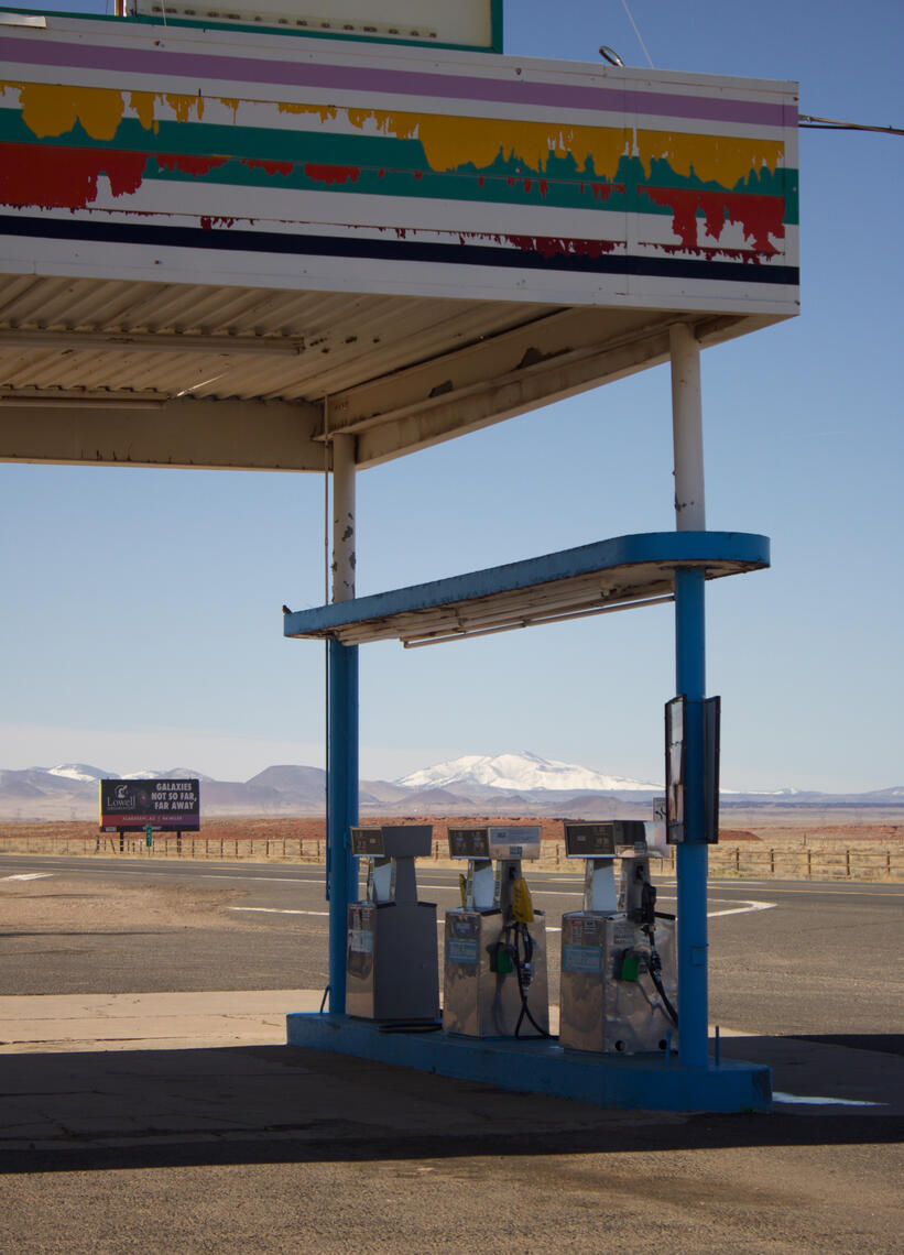 Old, weathered gas pumps under a covered shelter with a colorful, peeling sign above, set against a vast landscape with distant snow-capped mountains and a billboard.