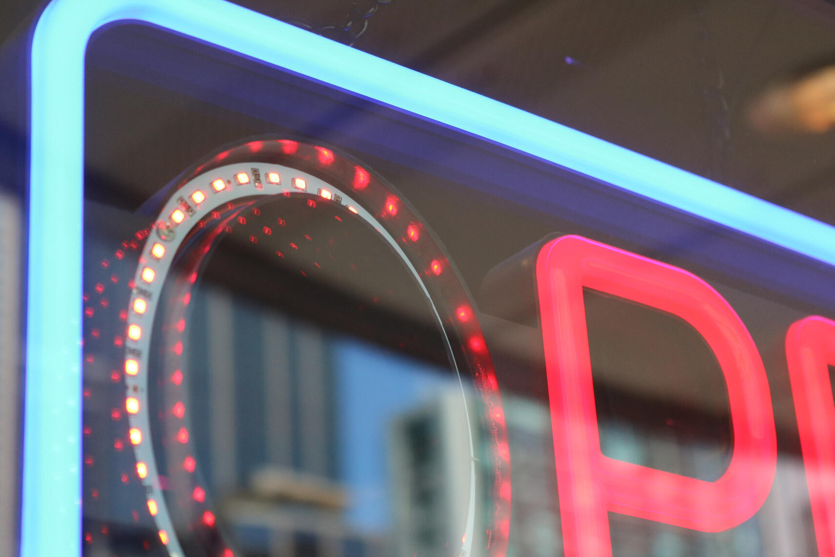 Close-up of a neon "Open" sign, featuring a bright red "P" and a circular ring of red LEDs in a glass window frame surrounded by blue neon tubing.