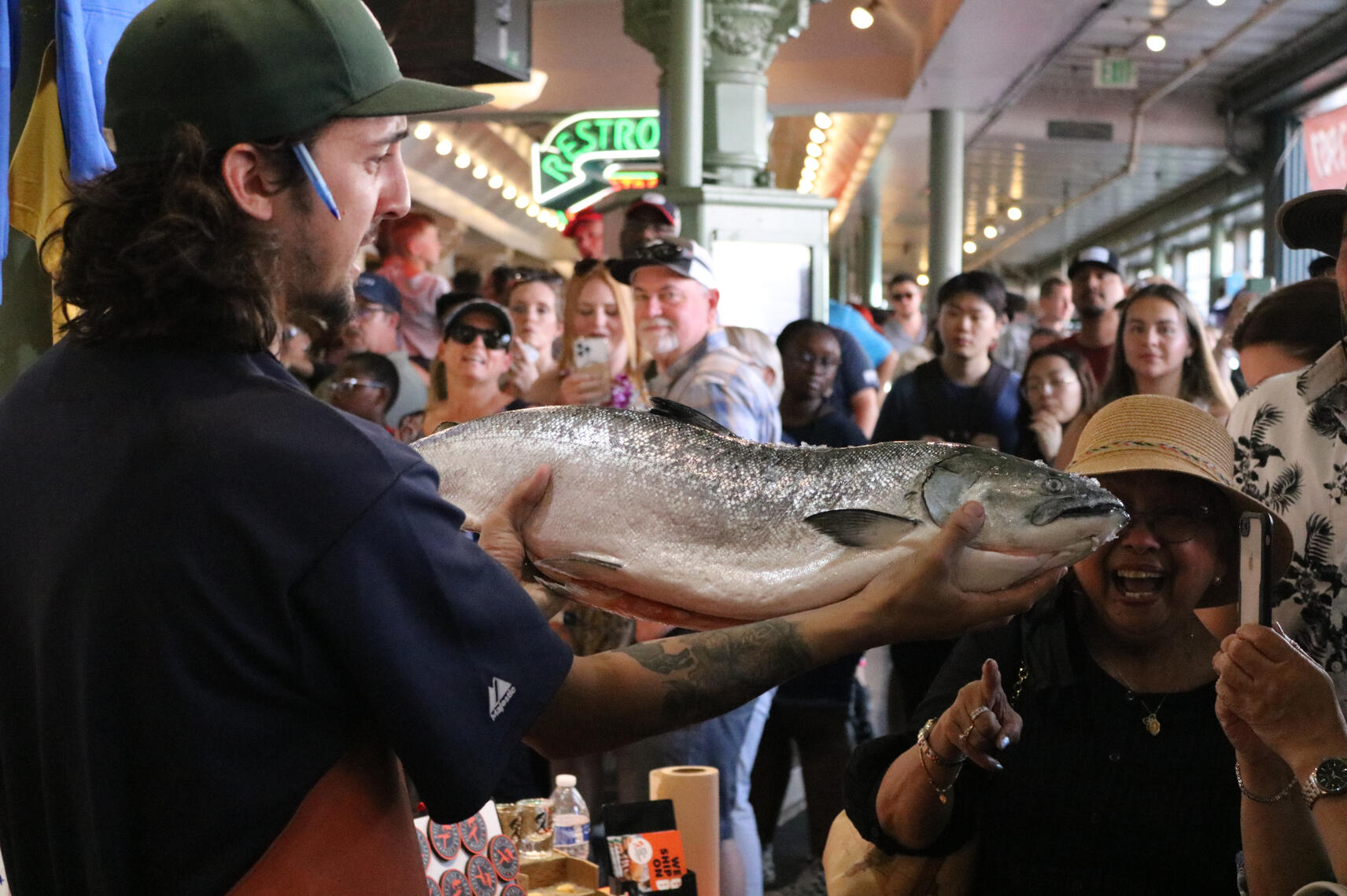 A man in a green baseball cap holds a large, freshly caught salmon aloft in a crowded marketplace, with an excited woman in a straw hat and several other onlookers smiling and taking photos.