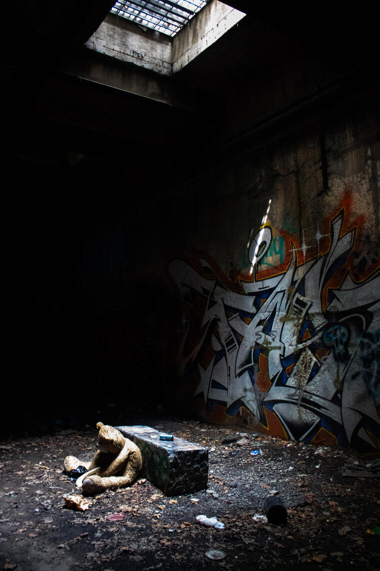 A worn teddy bear sits slumped against a concrete block in a dark, abandoned space, illuminated by a stark shaft of light from a skylight. Graffiti covers the concrete wall behind it.