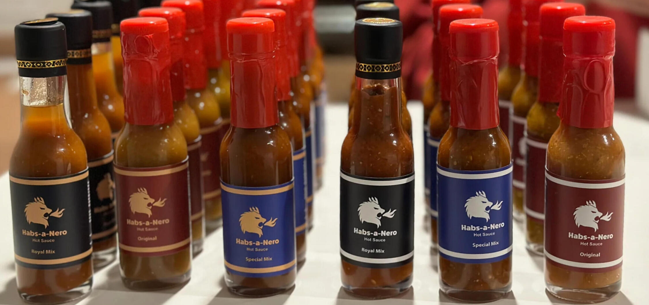 Rows of clear glass hot sauce bottles, labeled "Habs-a-Nero Hot Sauce". Some bottles have black labels, others have maroon or blue, and are sealed with red paper. The labels feature a gold illustration of a lion head breathing fire.