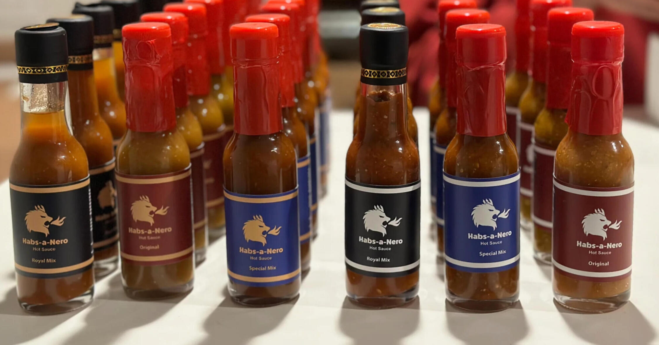 Rows of clear glass hot sauce bottles, labeled "Habs-a-Nero Hot Sauce". Some bottles have black labels, others have maroon or blue, and are sealed with red wax. The labels feature a gold illustration of a lion head breathing fire.