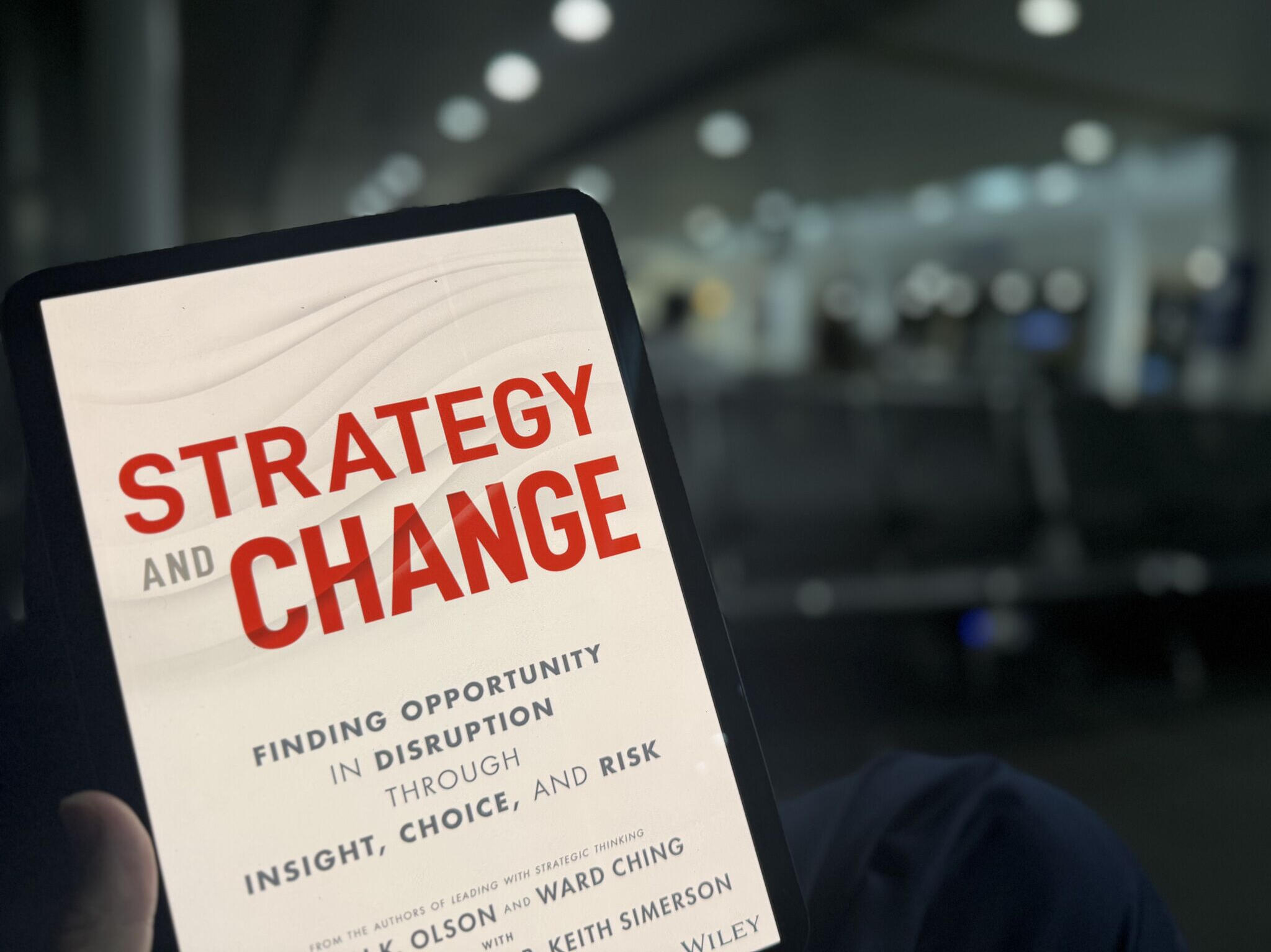 A close-up of a tablet or e-reader displaying the book cover titled “Strategy and Change” in large red letters, with the subtitle “Finding Opportunity in Disruption Through Insight, Choice, and Risk.” The device is held in the foreground, while a blurred a