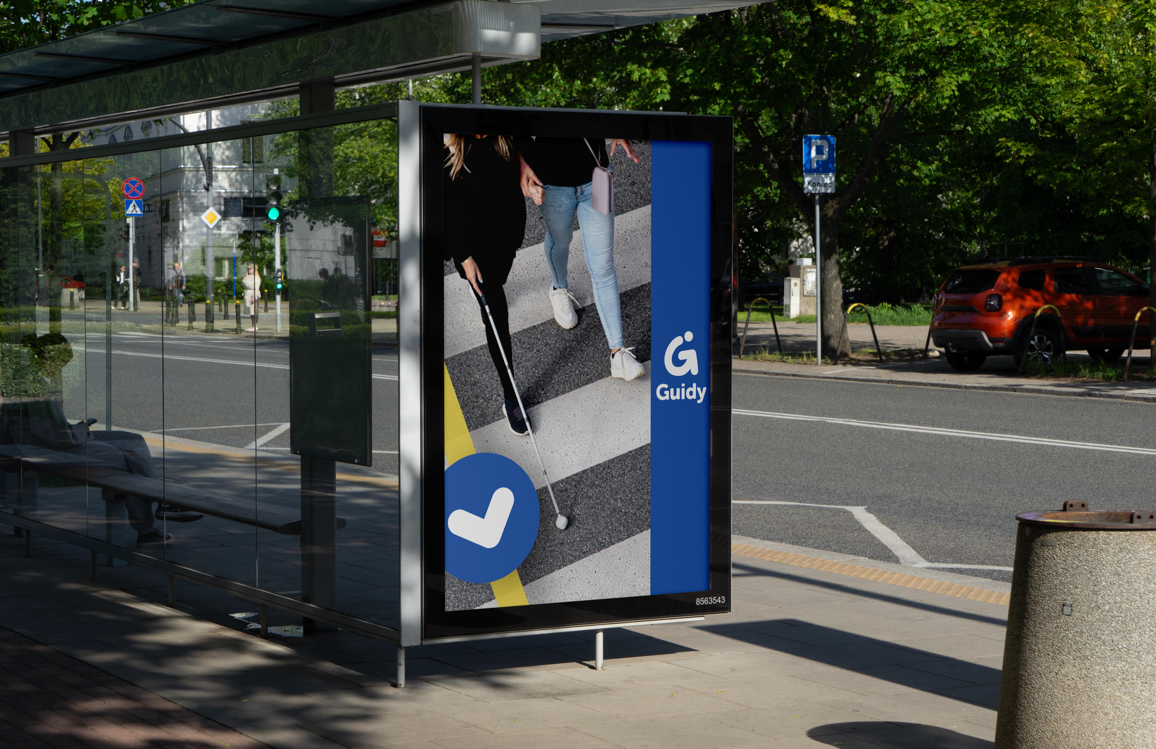An outdoor advertisement at a bus stop features two people crossing a crosswalk, one using a white cane, with a blue logo reading "Guidy" on the right. The bus stop is visible in the foreground, with trees and a road in the background.