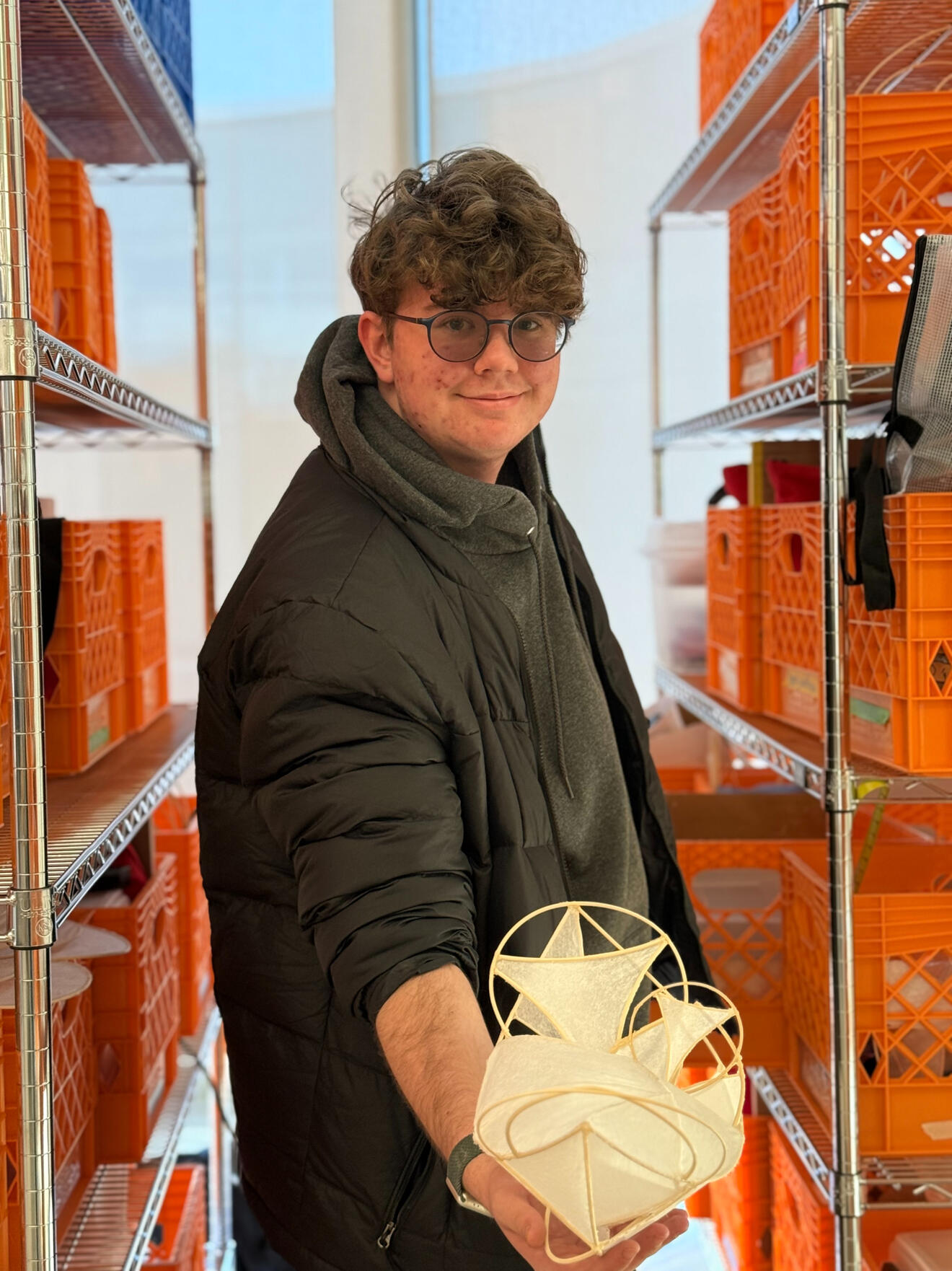 Liam in glasses and black jacket holding a small, geometric paper sculpture in front of a metal shelving unit filled with orange crates.